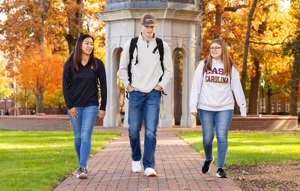 Three college students walking on a brick pathway through a campus area with autumn foliage and a stone gazebo in the background.