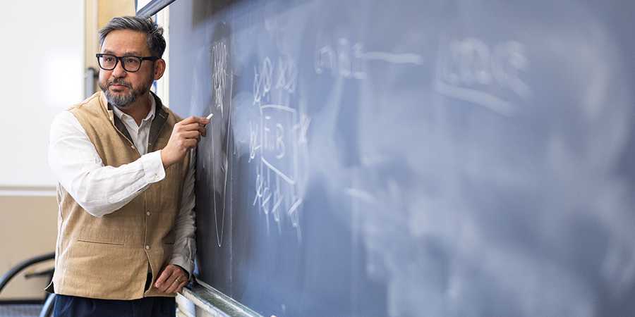 A male professor wearing glasses and a vest, writing on a large classroom chalkboard.