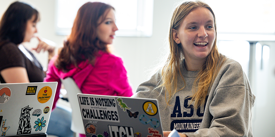 College students participating in a classroom discussion while using sticker-covered laptops.