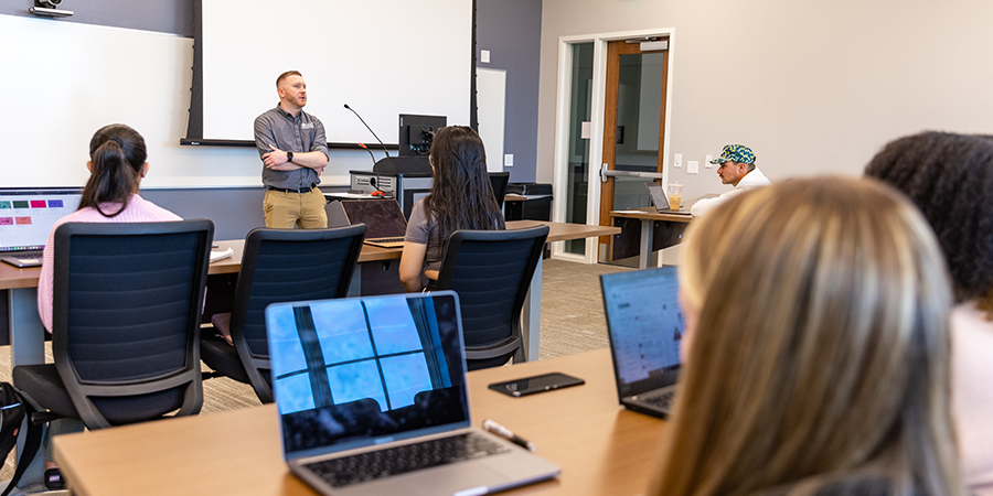 An instructor presenting to seated students in a classroom with laptops and a projection screen.