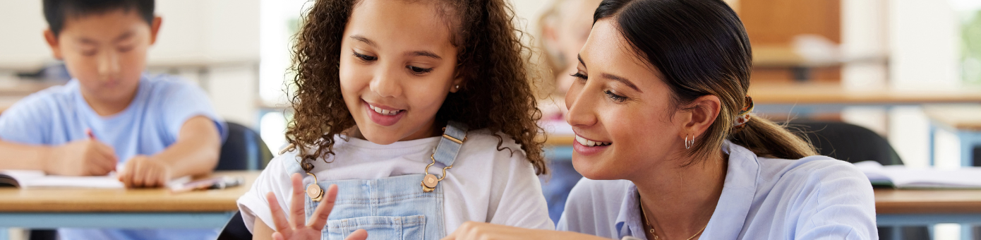 Young, female teacher smiles while helping a young girl with her school work in an elementary school classroom